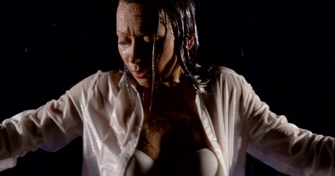 portrait of a dark-Haired girl in a white shirt and a white bra, she stands on a black background in the rain, dancing.