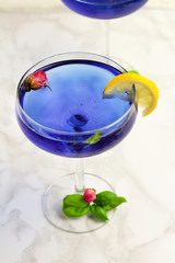 Thai asian blue Anchan tea in cocktail glasses with dry rosebuds, lemone slice and green basil leaves, healthy organic detox Asian herbal tea clitoria Butterfly Pea on marble background