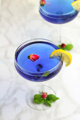 Thai asian blue Anchan tea in cocktail glasses with dry rosebuds, lemone slice and green basil leaves, healthy organic detox Asian herbal tea clitoria Butterfly Pea on marble background
