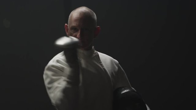 An older man fencer walks in a dark room and takes a bow with his foil