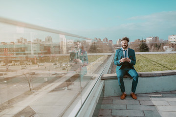 Fototapeta na wymiar Young smiling white collar worker in blue suit sitting on the rooftop with headphones on ears and using smart phone while looking at beautiful view.