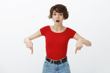 Surprised impressed cute pretty female student short haircut pointing down index fingers gasping drop jaw fascinationg astonishment stare camera excited stand white background amazement