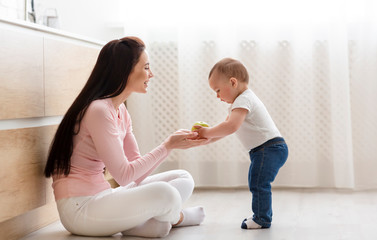 Mother giving fresh apple to baby, sitting on floor in kitchen