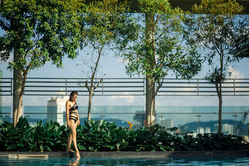Sexy slim caucasian brunette walking between green bushes and trees on edge of swimming pool on rooftop with cityscape © Kate