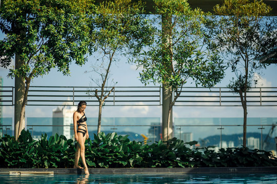 Sexy slim caucasian brunette walking between green bushes and trees on edge of swimming pool on rooftop with cityscape