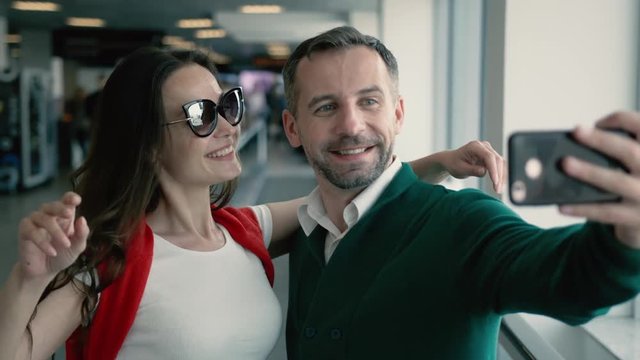 Young beautiful couple in the hall of airport. Brunette smiling woman and man taking selfie photo on smartphone near the window. Cinematic shot on RED camera.