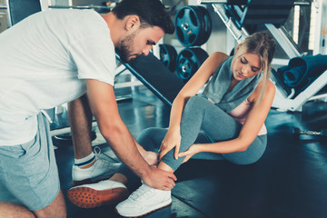 Pretty Woman Having Injury During Exercise in Gym While Her Couple Taking First Aid, Sporty Woman Injuries Painful Her Leg While Exercising. Accident and Sport Fitness Concept. - obrazy, fototapety, plakaty