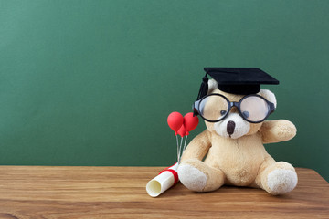 Teddy bear with graduation hat and diploma in front of green chalkboard. School concept