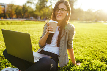 Image of beautiful stylish woman sitting on green grass with laptop  She is talking on the phone through wireless headphones. Sunset light. Lifestyle concept.