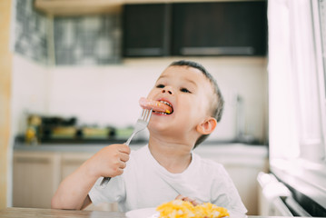 a child in a t-shirt in the kitchen eating a sausage and an omelet with a fork is very appetizing