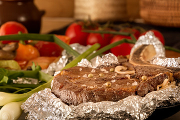 Meat steak baked in foil and baked vegetables on a wooden table with fresh tomatoes and greens. Close-up