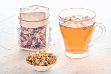 Herbal tea with pharmaceutical chamomile and a jar of dry rose petals on a white table. Close-up