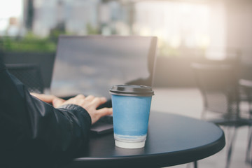 Paper cup of coffee and man hands on open laptop on the table of street cafe in business district of city