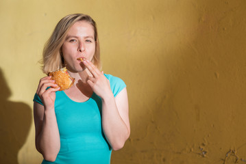 Portrait of a cute girl in a green dress walking outdoors and eating a burger on a warm summer day. Beautiful blonde is enjoying fast food at the yellow wall outside. Wrong food habits.