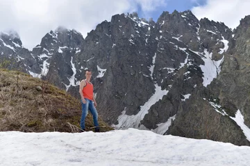 Fotobehang Athletic young man in the orange shirt on top of snow and the rocky mountains on a warm Sunny spring day. © Лариса Левина