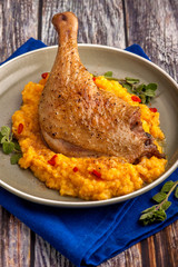 Roasted duck leg with pumpkin puree and fresh herbs. Large portion on a plate with a blue napkin on a wooden table. Close up and vertical view.