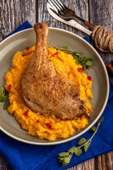 Roasted duck leg with pumpkin puree and fresh herbs. Large portion on a plate with a blue napkin on a wooden table. Close up and vertical view.
