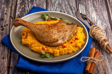 Roasted duck leg with pumpkin puree and fresh herbs. Large portion on a plate with a blue napkin on a wooden table. Close up and horizontal view.