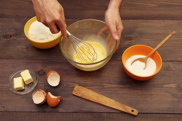 Chef hands knead dough with whisk in bowl on a wooden table.