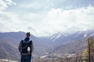 Rear view of a slim lonely young male traveler with a backpack reflexes and engaged in self-knowledge standing on the edge of a cliff overlooking a mountain forest valley