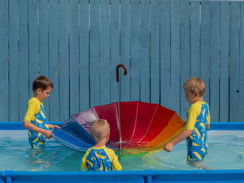 happy kids playing in carcas pool in boys' swimsuits with rainbow umbrella. brothers are happy together in warm water on sunny summer day. concept of educational games for preschoolers