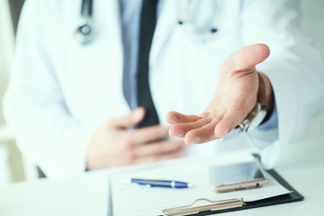 Male doctor making welcome gesture, politely inviting patient to sit down in medical office. Photo with depth of field.