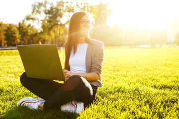 Image of beautiful stylish woman sitting on green grass with laptop  She is talking on the phone through wireless headphones. Sunset light. Lifestyle concept.