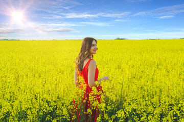 Young beautiful girl in a red dress close up in the middle of yellow field with radish flowers and sunlight..