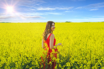 Naklejka na ściany i meble Young beautiful girl in a red dress close up in the middle of yellow field with radish flowers and sunlight..