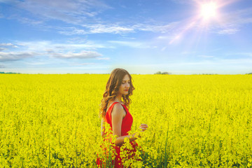 Fototapeta premium Young beautiful girl in a red dress close up in the middle of yellow field with radish flowers and sunlight..