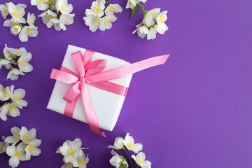 Gift with pink bow and white jasmine flowers on the violet background.Top view.Copy space.