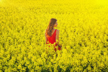 Young beautiful girl in a red dress close up in the middle of yellow field with radish flowers and sunlight..