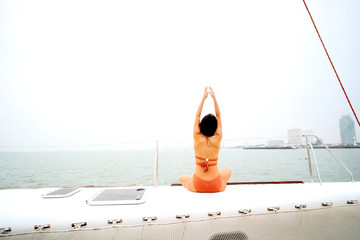 Back of young healthy and calm woman doing yoga meditation and sitting with hands up on sailing yacht boat with ocean sea in background