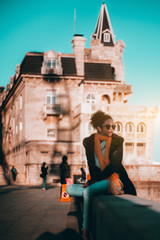 Young beautiful African-American female tourist is resting on a stone fence with an antique building behind; a vertical shot a biracial curly-hair girl in sunglasses sitting on the stone curb outdoors