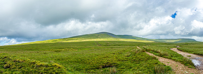 Obraz premium A panoramic view of a mountain with grassy green slope under a majestic blue sky and white clouds
