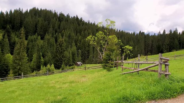 Picturesque spring mountains scene with wooden split rail fence across a green and lush pasture with a old house on the valley surrounded by pine woods.