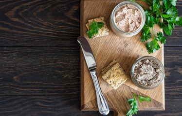 Fresh homemade liver pate in a glass jar with multigrain crackers and parsley on a wooden background.