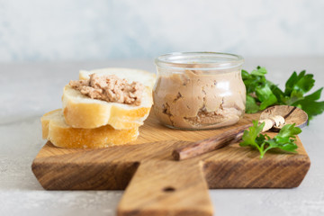 Liver pate in a glass jar with fresh bread and parsley on a wooden cutting board. Light grey stone background.