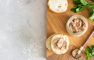 Liver pate in a glass jar with fresh bread and parsley on a wooden cutting board. Light grey stone background.