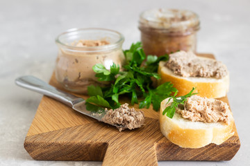 Liver pate in a glass jar with fresh bread and parsley on a wooden cutting board. Light grey stone background.