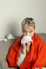 Unhealthy girl sitting on the couch, wrapped in a warm orange blanket, holding a napkin, sneezing...