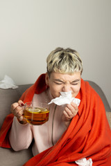 Unhealthy girl sitting on the couch, wrapped in a warm orange blanket, holding a napkin, sneezing and blowing her nose. Girl looks with chronic allergic rhinitis or cold difficult to breathe concept.