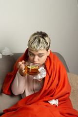 Unhealthy girl sitting on the couch, wrapped in a warm orange blanket, holding a napkin, sneezing...