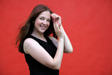 Photo portrait of a beautiful young pretty girl with dark red hair on a red background in a black jacket. Standing right in front of the camera in different poses, smiling, talking.