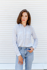 Young stylish beautiful brunette woman wearing blue striped shirt and denim jeans posing outdoors against white street wall. Trendy casual spring summer outfit. Street fashion.