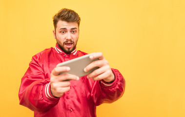 Portrait of an adult playing mobile games on a smartphone, focused looking at the screen, standing on a yellow background in a red jacket. Adult gamer plays mobile games, isolated.