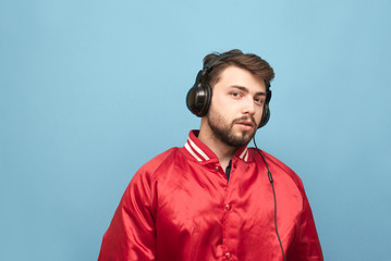 Portrait of a man with a beard, wearing a red jacket, stands on the background of a blue wall, and he listens to music in the headphones.Student with headphones isolated.