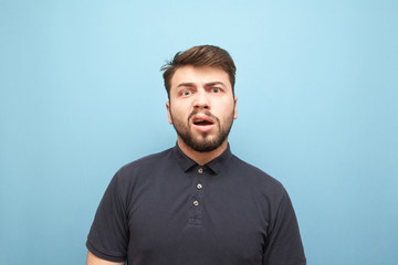 Close-up portrait of an emotional man with a beard, looks at the camera with astonished face and is angry.Funny man in a dark shirt with an evil face on a blue background. Isolated.Man is in shock