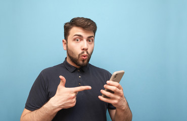 Amazed man in a dark T-shirt and beard, holds a smartphone in his hands, looks emotionally in the camera and shows the index finger on the phone. Emotional man with a smartphone in his hand. Isolated.