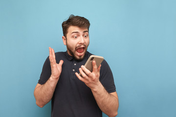 Portrait of a crazy bearded man with a smartphone in his hands, looking at the screen and screaming on a blue background. Shocked man uses a smartphone and rejoices. Isolated.